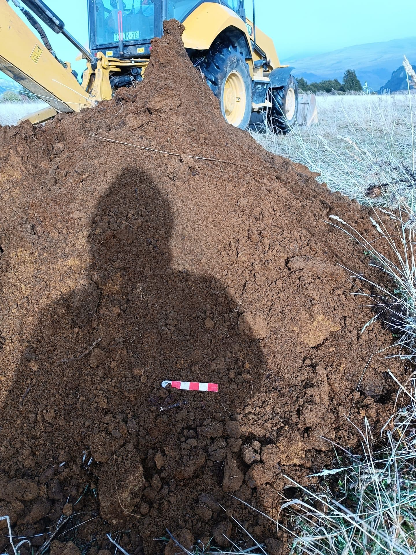 Calicata con escala de medición junto a una excavadora en terreno.